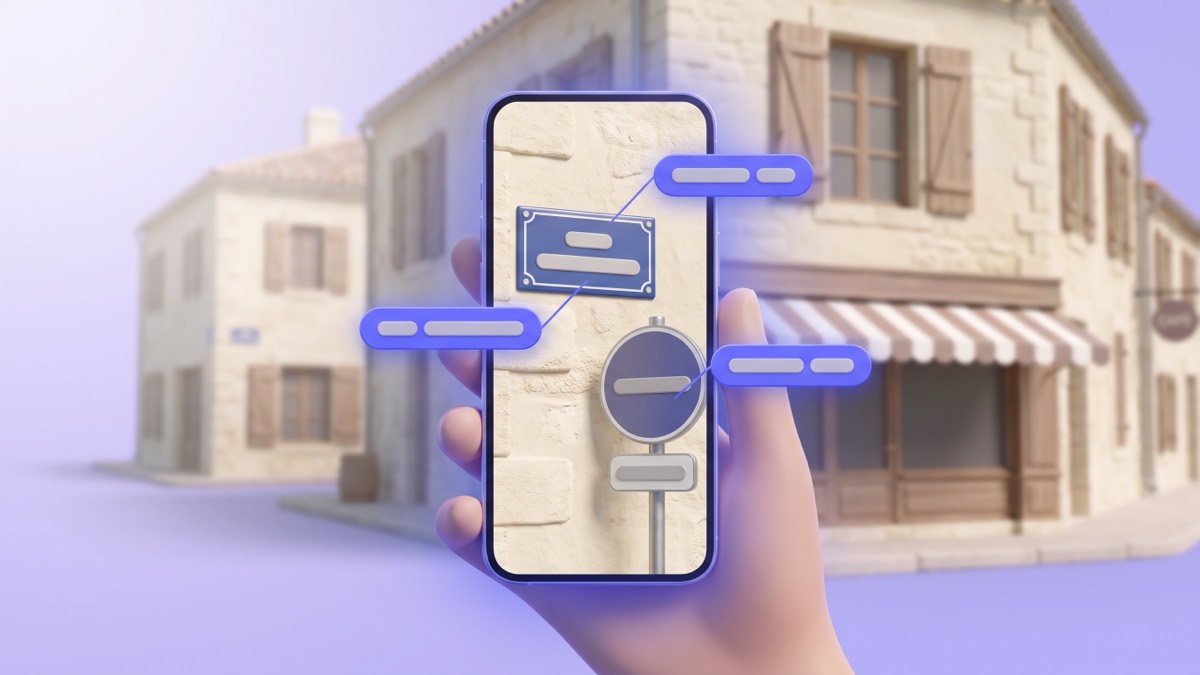 A phone held up on a quiet French village street, its screen showing a blue street name plaque and a road sign with indigo translation chips over them, with cream stone houses and a café awning behind